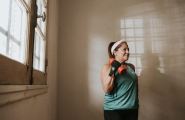 Senior woman in a brightly lit room exercises with dumbbells, promoting fitness at home.