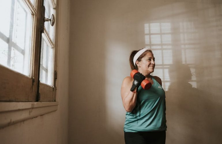 Senior woman in a brightly lit room exercises with dumbbells, promoting fitness at home.